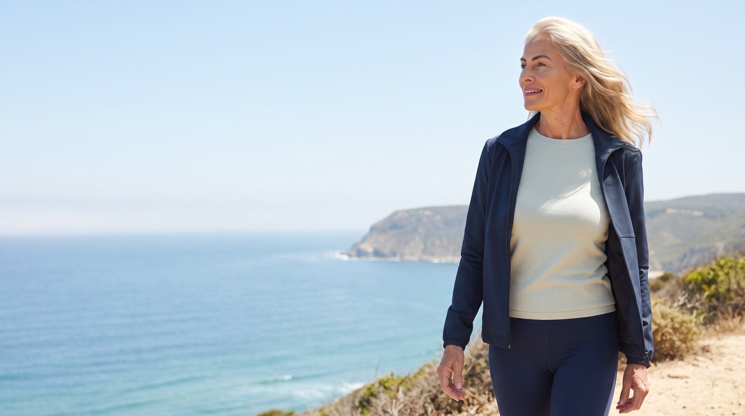 A woman in her fifties walking a clifftop coastal path on a bright clear morning, the ocean open beside her